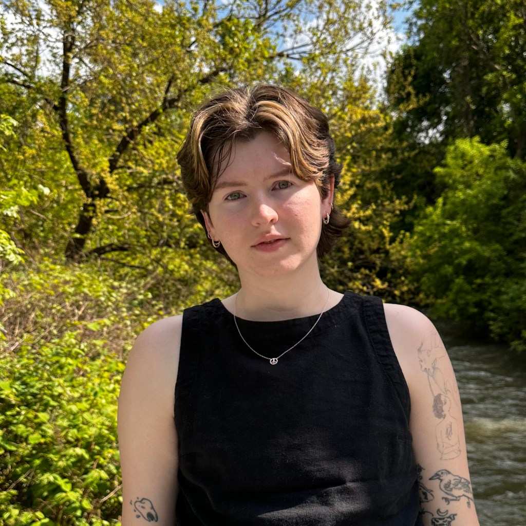 a photo of the author, macks cook, a white, tattooed nonbinary person with brown hair, standing in nature with a creek in the background